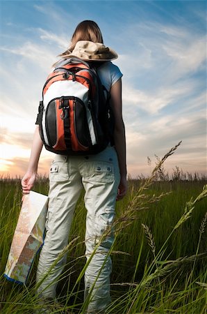 beautiful young woman with backpack and map in hand standing outside in the field with her back to camera. Sunset cloudy blue sky in background and green grass in foreground Photographie de stock - Aubaine LD & Abonnement, Code: 400-04200705