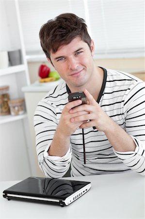 simsearch:400-04212823,k - Smiling man sending a text message with his cellphone in front of his laptop in the kitchen at home Stock Photo - Budget Royalty-Free & Subscription, Code: 400-04341573