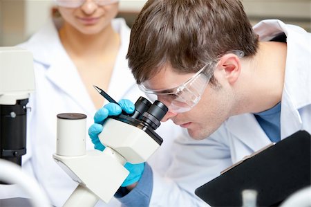 simsearch:400-05669968,k - Caucasian male scientist holding pen and clipboard looking through a microscope in his laboratory Stock Photo - Budget Royalty-Free & Subscription, Code: 400-04341763