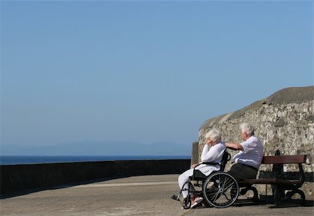 Elderly woman in a wheelchair with her hand on her head, seemingly depressed sitting next to an elderly man. Sea view and a blue sky in the background. Stock Photo - Budget Royalty-Free & Subscription, Code: 400-04945150