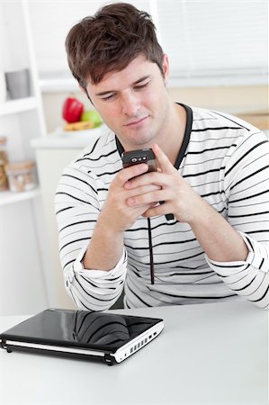 simsearch:400-04212823,k - Caucasian man sending a text message with his cellphone in front of his laptop in the kitchen at home Stock Photo - Budget Royalty-Free & Subscription, Code: 400-05320820