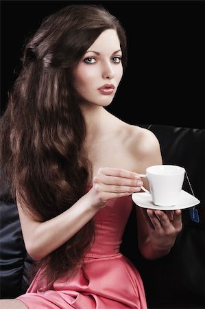 sophisticated young pretty brunette in pink elegant dress sitting on a black sofa and tasting a cup of tea. she looks in to the lens with  three quarters turned faces  and takes a cup of tea with both hands Stock Photo - Budget Royalty-Free & Subscription, Code: 400-05752152