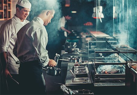 Food concept. Chef in white uniform monitors the degree of roasting and greases meat with oil in saucepan in interior of modern restaurant kitchen. Preparing traditional beef steak on barbecue oven. Photographie de stock - Aubaine LD & Abonnement, Code: 400-09028536