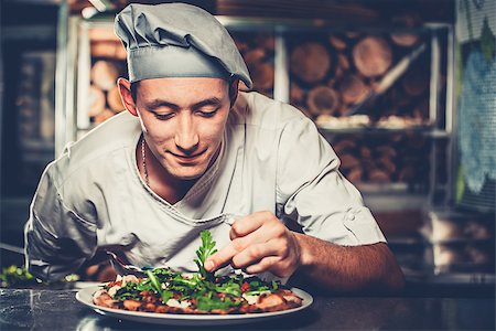 Food concept. Preparing traditional italian pizza. Young smiling chef in white uniform and gray hat decorate ready dish with green rucola herbs in interior of modern restaurant kitchen. Ready to eat. Stock Photo - Budget Royalty-Free & Subscription, Code: 400-09028539