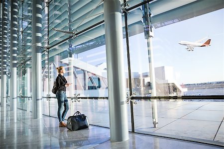 simsearch:695-05773315,k - Young casual female traveler at airport, holding smart phone device, looking through the airport gate windows at planes on airport runway. Stock Photo - Budget Royalty-Free & Subscription, Code: 400-09085017