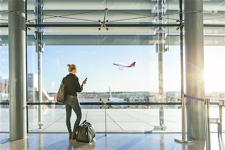 simsearch:695-05773315,k - Young casual female traveler at airport, holding smart phone device, looking through the airport gate windows at planes on airport runway. Stock Photo - Budget Royalty-Free & Subscription, Code: 400-09170476