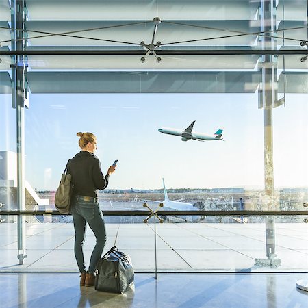 simsearch:695-05773315,k - Young casual female traveler at airport, holding smart phone device, looking through the airport gate windows at planes on airport runway. Stock Photo - Budget Royalty-Free & Subscription, Code: 400-09170477