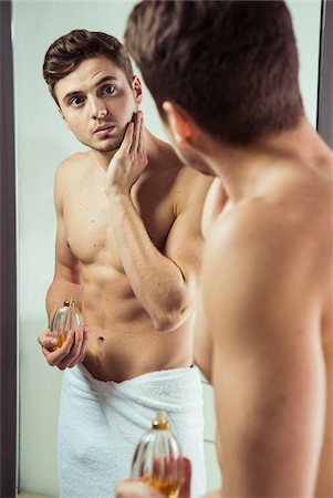 Young man looking in bathroom mirror with towel wrapped around waist, applying cologne to face, studio shot Stock Photo - Premium Royalty-Free, Code: 600-07278949