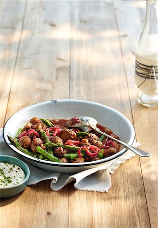 Metal bowl filled with baked mushrooms, asparagus, and red onion in a tomato sauce with cracked black pepper. Sour cream sauce with fresh chives shown on the side. Meal shown with a spoon on a wooden tabletop, studio shot Stock Photo - Premium Royalty-Free, Code: 600-07311137