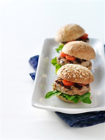 Three grilled turkey burger sliders on whole wheat buns with tomato and lettuce on a white platter in a high key table top setting with a blue cloth napkin. Stock Photo - Premium Royalty-Free, Code: 600-07311154