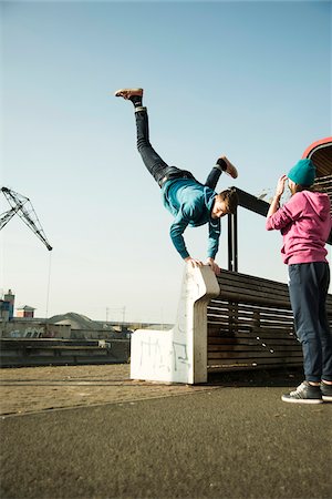 Teenage boy doing handstand on bench outdoors, while teenage girl watches, industrial area, Mannheim, Germany Foto de stock - Sin royalties Premium, Código: 600-07584770