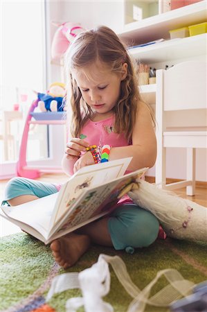 Little Girl Sitting on the Floor of her Bedroom, Reading a Book and Looking at her Bracelet Stock Photo - Premium Royalty-Free, Code: 600-07743454