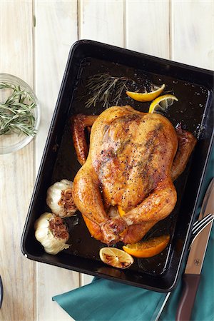 Roast chicken, garlic, rosemary and lemon in a roasting pan with a carving knife and fork, studio shot Photographie de stock - Premium Libres de Droits, Code: 600-08002118