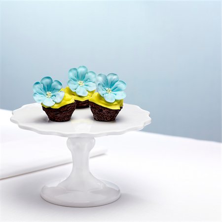 Chocolate Cupcakes with Yellow Icing and Blue Sugar Flowers on Cake Stand Stock Photo - Premium Royalty-Free, Code: 600-08512618