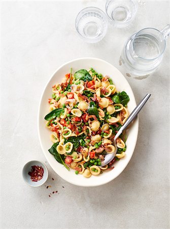 Platter of orecchiette pasta with peas, bacon, spinach and tomatoes with chili flakes in a small bowl and water glasses and pitcher on a beige background Photographie de stock - Premium Libres de Droits, Code: 600-09155580