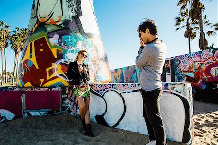 Young woman standing by graffiti art at beach, young man photographing her Stock Photo - Premium Royalty-Free, Code: 614-08544726