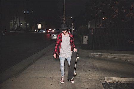 Male hipster skateboarder walking along sidewalk at night Stock Photo - Premium Royalty-Free, Code: 614-08877269