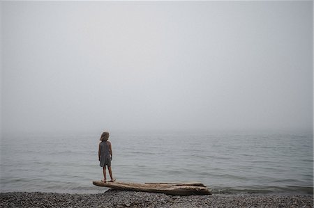 Girl standing on driftwood looking out to sea Stock Photo - Premium Royalty-Free, Code: 614-09057054
