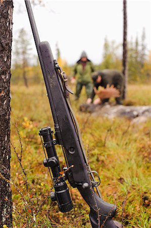 simsearch:6102-06337050,k - Rifle leaning on tree trunk, hunters in background Stock Photo - Premium Royalty-Free, Code: 6102-08760193