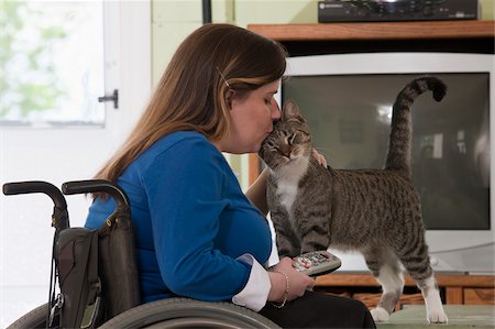 disable - Woman with Spina Bifida in a wheelchair kissing a cat while holding the TV remote Stock Photo - Premium Royalty-Free, Code: 6105-05397276