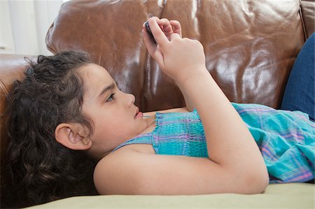 sending - Hispanic girl lying on a couch and looking at a text message on a mobile phone Foto de stock - Sin royalties Premium, Código: 6105-05397215
