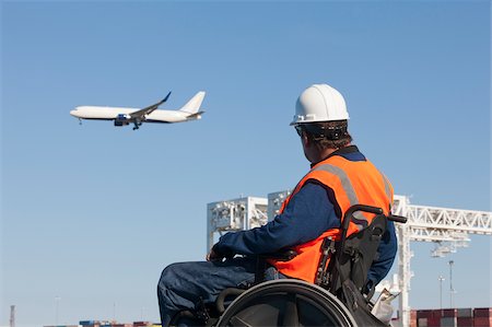 plane and sky - Transportation engineer in wheelchair watching airliner flying over shipping containers and cranes at port Stock Photo - Premium Royalty-Free, Code: 6105-05953720