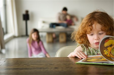 Little girl reading children's book, family in the background Stock Photo - Premium Royalty-Free, Code: 6108-05856631