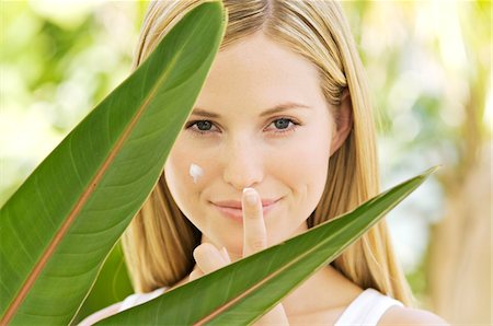 simsearch:6108-05857519,k - Portrait of a young woman looking at the camera, applying moisturizer on her face, house plant leaves, outdoors Stock Photo - Premium Royalty-Free, Code: 6108-05857525