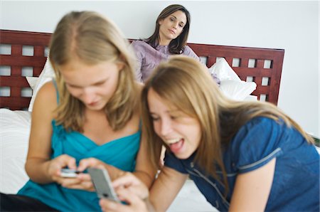 family indoors summer - 2 smiling teenage girls using mobile phones, woman in background Stock Photo - Premium Royalty-Free, Code: 6108-05857805