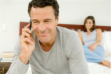 simsearch:6108-05857751,k - Portrait of a man phoning with cell, woman sitting on bed in background, indoors Stock Photo - Premium Royalty-Free, Code: 6108-05858309