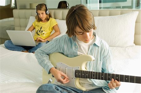 Boy playing a guitar and girl working on a laptop behind him Stock Photo - Premium Royalty-Free, Code: 6108-05862977