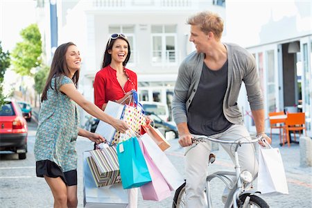 Young man cycling with two women running after him with shopping bags Stock Photo - Premium Royalty-Free, Code: 6108-05869578