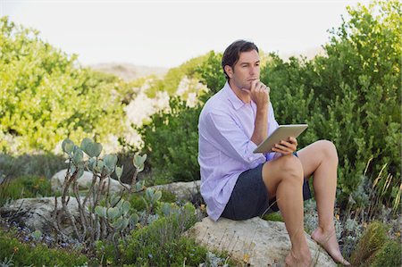 Man sitting on a rock using a digital tablet and thinking Stock Photo - Premium Royalty-Free, Code: 6108-05869659