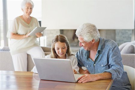 Man using a laptop with his granddaughter and his wife working on a digital tablet in background Stock Photo - Premium Royalty-Free, Code: 6108-05870137