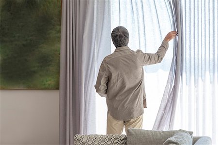 Rear view of a man holding curtain on window Stock Photo - Premium Royalty-Free, Code: 6108-08663333