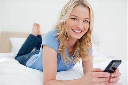 A woman lying on the bed as she smiles looking straight ahead, as she uses her smartphone. Stock Photo - Premium Royalty-Free, Code: 6109-06003043