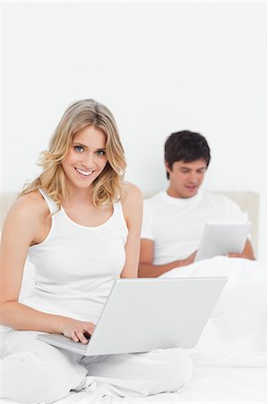 A man using the tablet and woman using the laptop are both smiling while sitting on the bed. Stock Photo - Premium Royalty-Free, Code: 6109-06003255