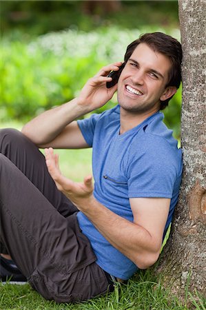 Jeune homme heureux, à l'aide de son téléphone portable tout en penchant contre un arbre dans un parc Photographie de stock - Premium Libres de Droits, Code: 6109-06003479