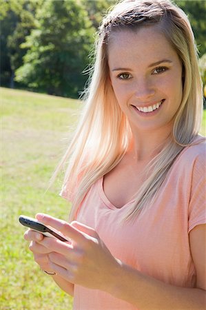 Young smiling blonde girl looking at the camera while sending a text in a park Stock Photo - Premium Royalty-Free, Code: 6109-06003563