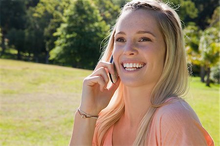 Young smiling blonde girl standing up in a parkland while using her mobile phone Stock Photo - Premium Royalty-Free, Code: 6109-06003561