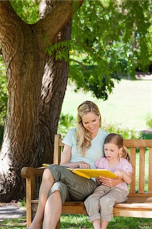 A close up shot of a smiling kid and her mom reading a book together as they sit on the bench Stock Photo - Premium Royalty-Free, Code: 6109-06003946