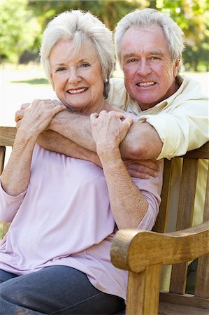 Man and woman smiling and looking to their left side while he is holding her from behind the bench Stock Photo - Premium Royalty-Free, Code: 6109-06004623