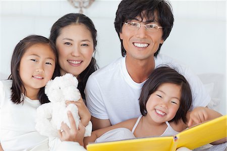 A smiling family sits on the bed together as they hold a book and a teddy bear Stock Photo - Premium Royalty-Free, Code: 6109-06005015