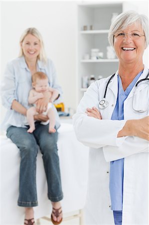 doctor and child patient and mom - Smiling pediatrician with arms folded and mother with her little baby behind her Stock Photo - Premium Royalty-Free, Code: 6109-06006567