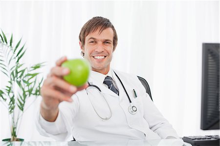 Smiling surgeon holding a green apple while sitting at the desk behind his computer Stock Photo - Premium Royalty-Free, Code: 6109-06006867