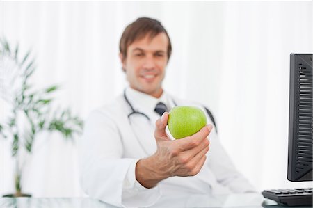 Green apple being held by a smiling surgeon sitting at his desk Stock Photo - Premium Royalty-Free, Code: 6109-06006864