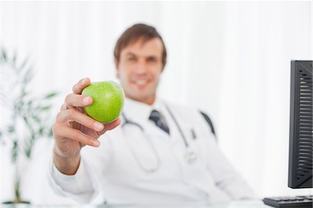Green apple being held by a relaxed doctor sitting behind his computer Stock Photo - Premium Royalty-Free, Code: 6109-06006866