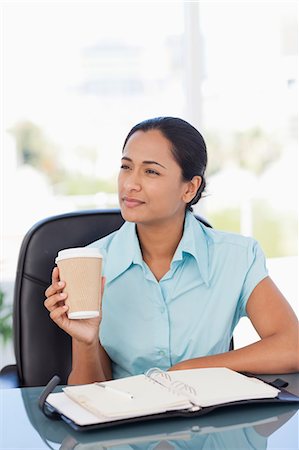 Young secretary holding a coffee while sitting at a desk and looking towards the side Stock Photo - Premium Royalty-Free, Code: 6109-06006976