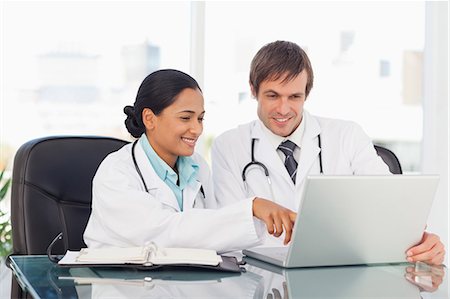 Smiling doctors talking while sitting at the desk in front of a laptop and a clipboard Stock Photo - Premium Royalty-Free, Code: 6109-06006996