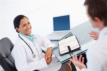 Happy doctor listening to a co-worker while sitting at a desk with a diary Stock Photo - Premium Royalty-Free, Code: 6109-06007015